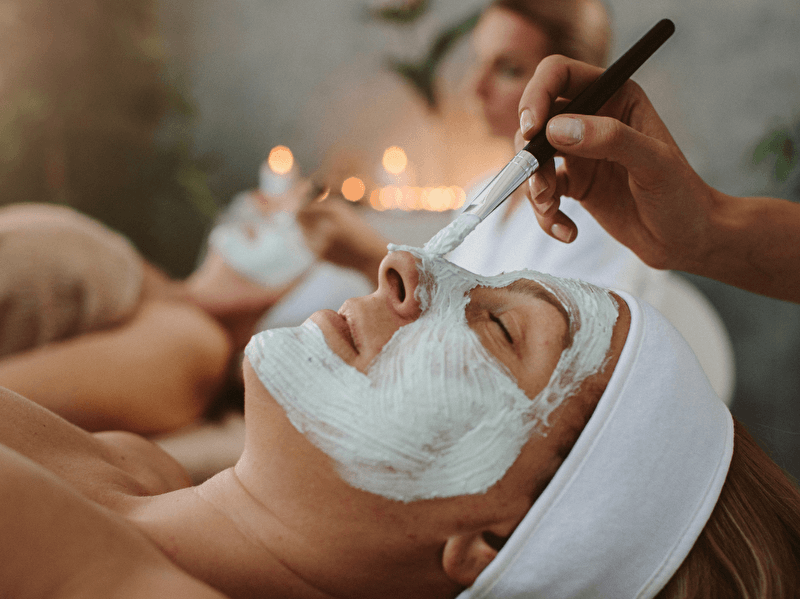 Vrouw ontvangt ontspannende gezichtsbehandeling met een verzorgend masker in een sfeervolle spa-omgeving, met zachte verlichting en kaarsjes op de achtergrond.